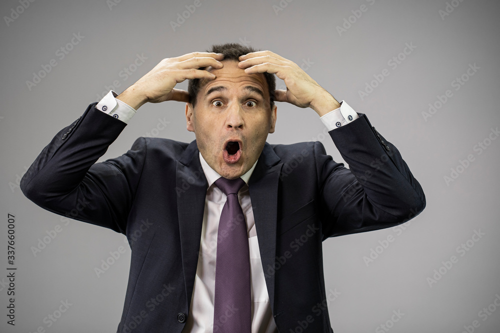 Shocked middle age businessman in formal clothes with hands on head feeling worried about deadlines, falling stocks, company delivery, closing, bankruptcy concept. Isolated on grey background
