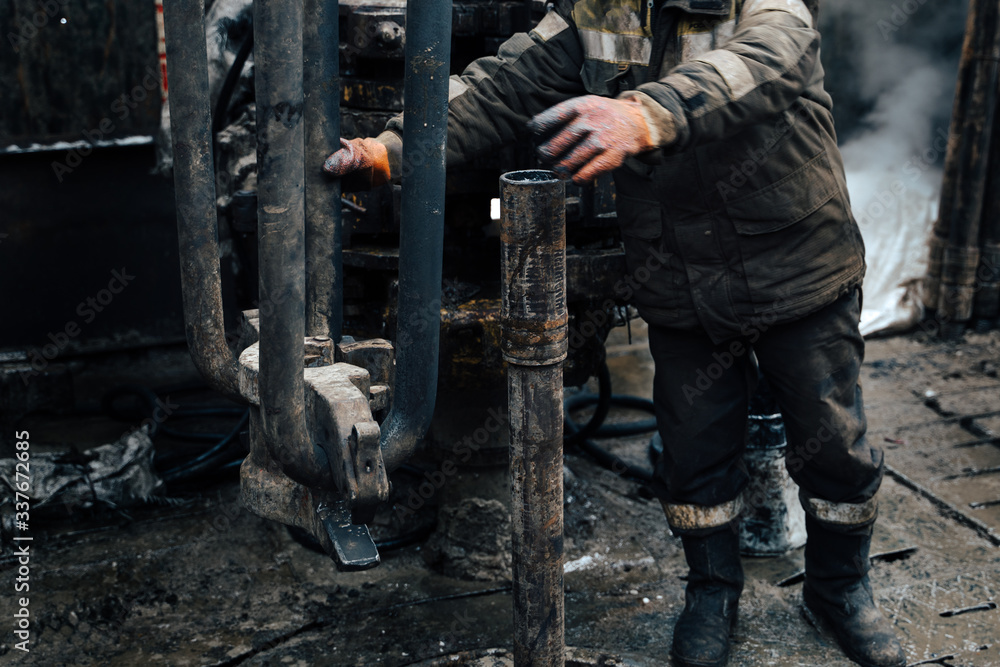 Work driller in red uniform, in helmet and goggles. He uses a hydraulic wrench to screw drill pipes to lower them into an oil well and continue drilling it. The concept of a working person. Work driller in red uniform, in helmet and goggles. He uses a hydraulic wrench to screw drill pipes to lower them into an oil well and continue drilling it. The concept of a working person.