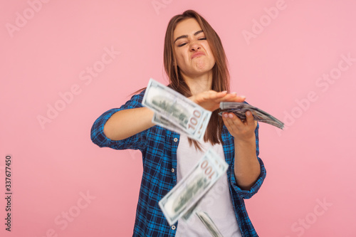 Portrait of wasteful rich girl in checkered shirt scattering dollars with arrogant grimace, boasting wealthy life, concept of careless money spending. indoor studio shot isolated on pink background