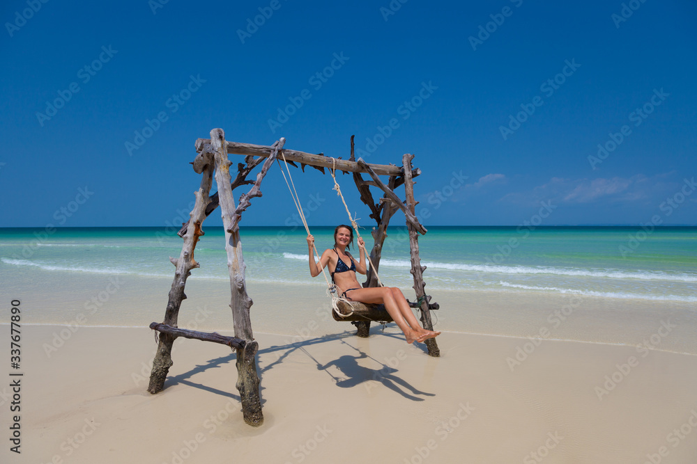 Junge fröhliche Frau, die auf einer Schaukel im türkisblauen Wasser an einem Paradiesstrand entspannt und in einem tropischen Land Urlaub macht