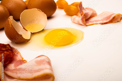 Broken chicken egg  scattered chicken eggs  slices of sausage and bacon and tomatoes on a white background. Close up.