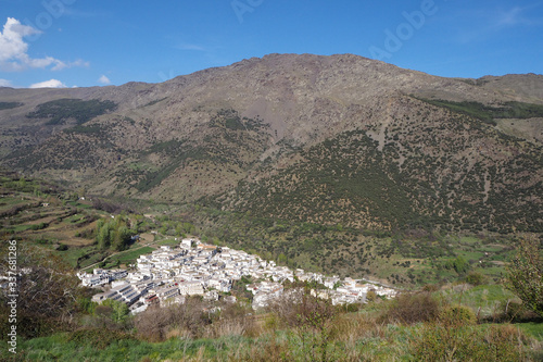 The mountain landscape with the slope covered by green trees and shrubs, the far village with white houses in the beautiful valley on the sunny spring day.