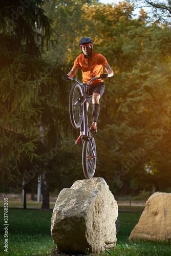 Portrait of young cyclist standing on back wheel of bmx bike on a rock looking at camera in park, green trees on background photo