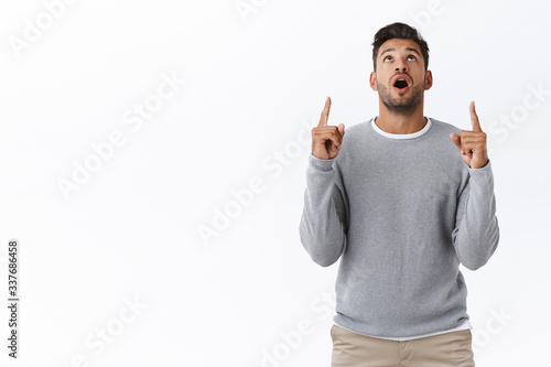 Surprised, wondered handsome bearded guy with stylish haircut, wear casual grey sweater, drop jaw impressed, raise head and staring upwards from amazement, pointing up with interest and curiosity