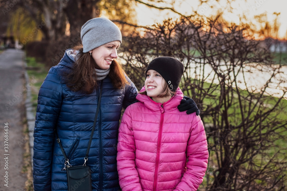 Fototapeta premium A woman and her daughter are walking by the river and talking, smiling at each other, the girl began her teenage period and her mother helps her to overcome depression