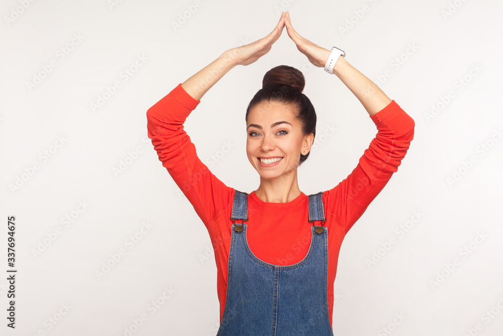 I'm in safety. Portrait of happy stylish pretty girl with hair bun in denim overalls standing with roof gesture and smiling contentedly, dreaming of house. studio shot isolated on white background