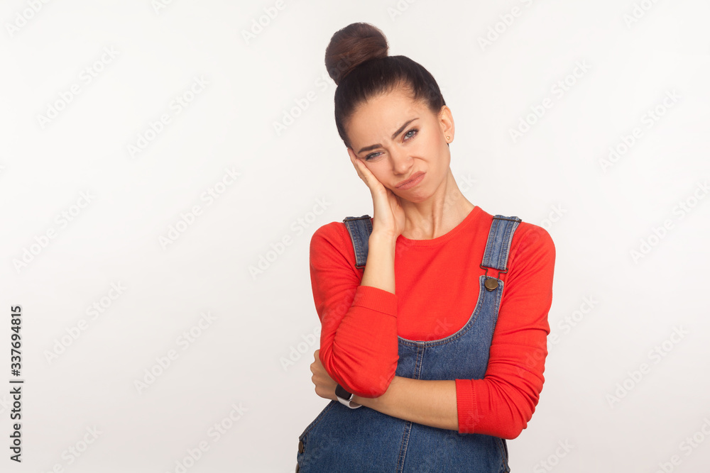 Portrait of frustrated indifferent girl with hair bun in denim overalls leaning on hand expressing disinterest to tedious boring talk, lack of energy, depression. indoor studio shot white background Portrait of frustrated indifferent girl with hair bun in denim overalls leaning on hand expressing disinterest to tedious boring talk, lack of energy, depression. indoor studio shot white background