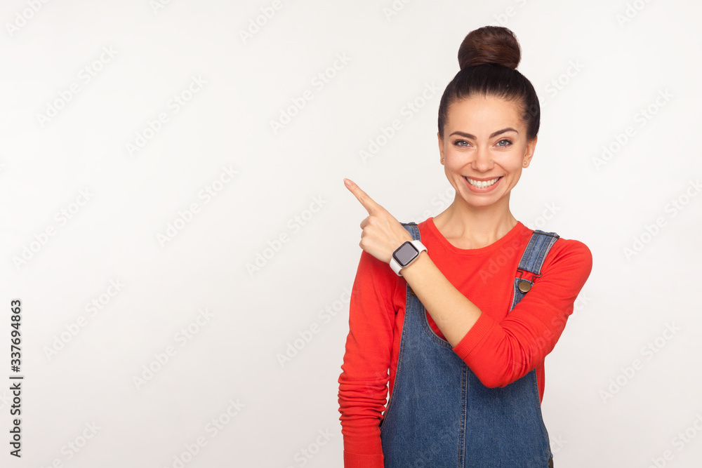 Attention to advertising! Portrait of happy stylish pretty girl with hair bun in denim overalls pointing aside to copy space, blank wall for commercial text. studio shot isolated on white background Attention to advertising! Portrait of happy stylish pretty girl with hair bun in denim overalls pointing aside to copy space, blank wall for commercial text. studio shot isolated on white background