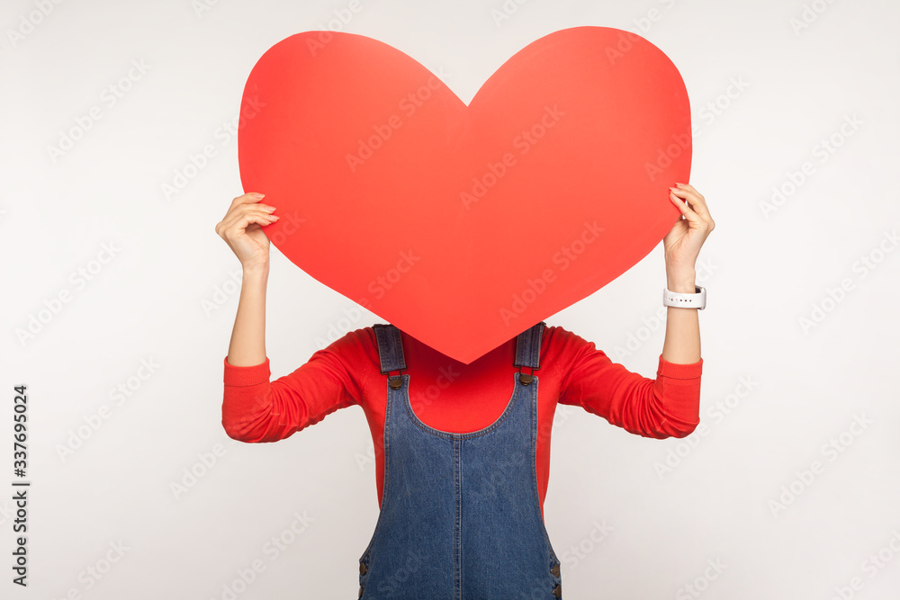 Portrait of anonymous girl in denim overalls hiding face behind big red  paper heart, holding huge greeting card on Valentine's day, expressing love  and romance. indoor studio shot, white background Photos
