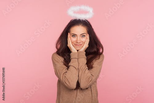 Portrait of modest young pretty woman with halo above head holding hands on face and looking at camera with kind timid smile, angelic beauty, shyness. indoor studio shot isolated on pink background