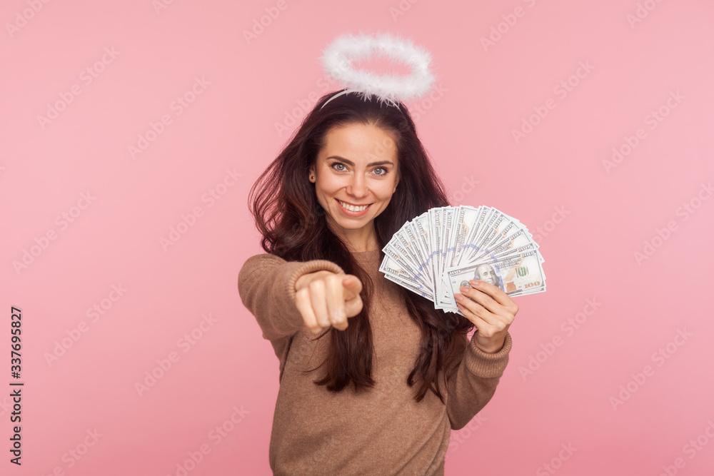 Portrait of happy angelic young woman with halo above head holding dollar banknotes and pointing to camera, choosing lottery winner, encouraging to earn big money. indoor studio shot, pink background Portrait of happy angelic young woman with halo above head holding dollar banknotes and pointing to camera, choosing lottery winner, encouraging to earn big money. indoor studio shot, pink background