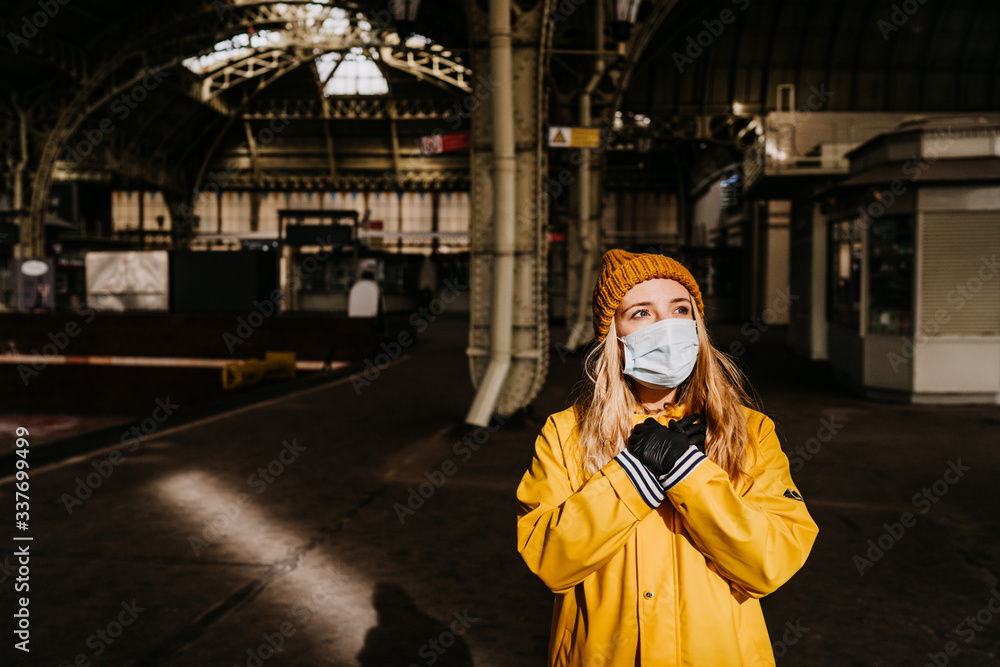 Blonde in medical protective mask, yellow windbreaker stands alone at the train station, looking at the sun and waiting for a good news, concerned about coronavirus pandemic. Covid-19 virus concept. Blonde in medical protective mask, yellow windbreaker stands alone at the train station, looking at the sun and waiting for a good news, concerned about coronavirus pandemic. Covid-19 virus concept.