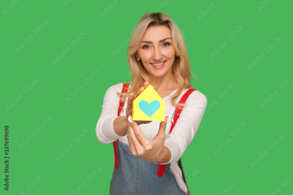 Home purchase, real estate insurance. Portrait of happy beautiful adult blond woman in urban style overalls showing paper house with heart inside and smiling. studio shot isolated on green background Home purchase, real estate insurance. Portrait of happy beautiful adult blond woman in urban style overalls showing paper house with heart inside and smiling. studio shot isolated on green background