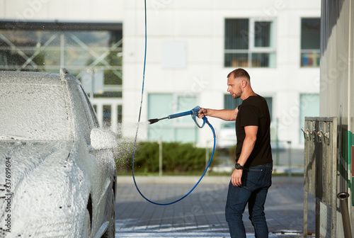 Man in dark clothes is washing his car holding a hose with foam outdoors, side view