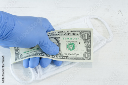 Ludzka ręka w niebieskiej rękawicy ochronnej trzymającej pakiet banknotów i maskę ochronną.