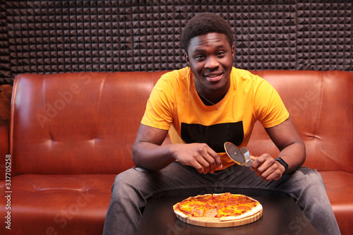 A black young man in a yellow t-shirt is sitting on a leather sofa with a pizza knife in his hand. Photo in the interior. Copy of the space. photo