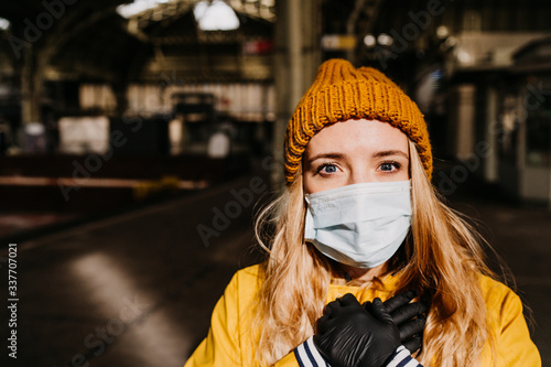 Close up portrait of scared blonde in protective medical mask, protective gloves and yellow jacket at empty railway station, she is very upset about the coronavirus. Covid-19 virus concept.. © iona
