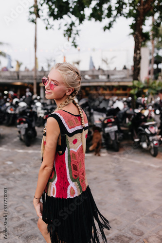 Portrait from back of pleasant white girl in stylish knitted attire. Outdoor shot of cute fair-haired female model with braids posing on blur background with gently smile.