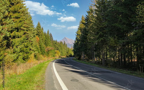 Country asphalt road, coniferous trees on both sides, mount Krivan peak (Slovak symbol) with blue sky above, in distance
