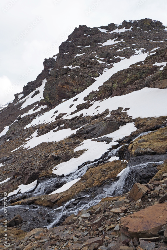 The gloomy mountain landscape with the dark rocks and stones, the waterfall, the slope covered by white snow on the cloudy day.