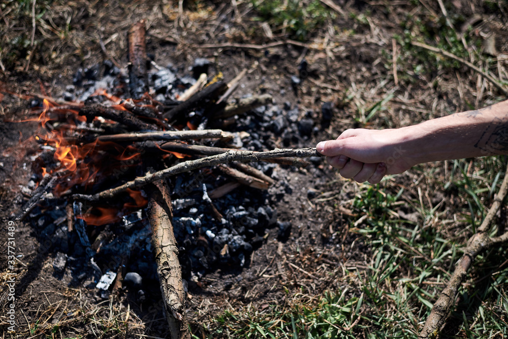 Ternopil/Ukraine-Apr 04 2020: Close-up picture of bonfire, made of dry branches, outside on green grass at sunny weather in summer. Hand, holding wooden stick. fixing fireplace in forest.