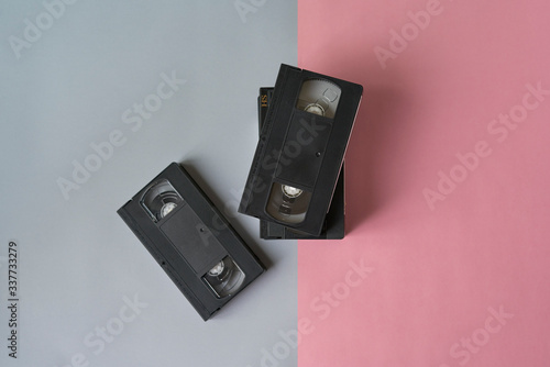 Pile of black vhs videotapes on a pink and grey background.