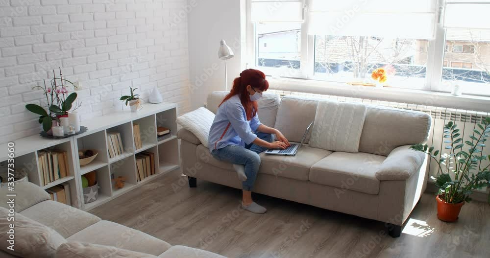 Work from home during COVID-19. Businesswoman with a medical mask working from home on a laptop, focused girl using computer sitting on a couch, female user busy on distance internet job, 4k Work from home during COVID-19. Businesswoman with a medical mask working from home on a laptop, focused girl using computer sitting on a couch, female user busy on distance internet job, 4k