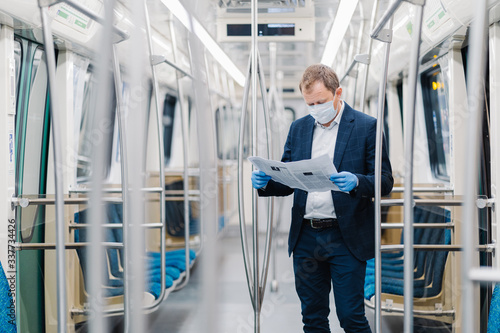 Young male entrepreneur wears disposable medical mask and gloves in metro, prevents coronavirus disease, poses in empty carriage, reads newspaper, finds out information about contagious virus