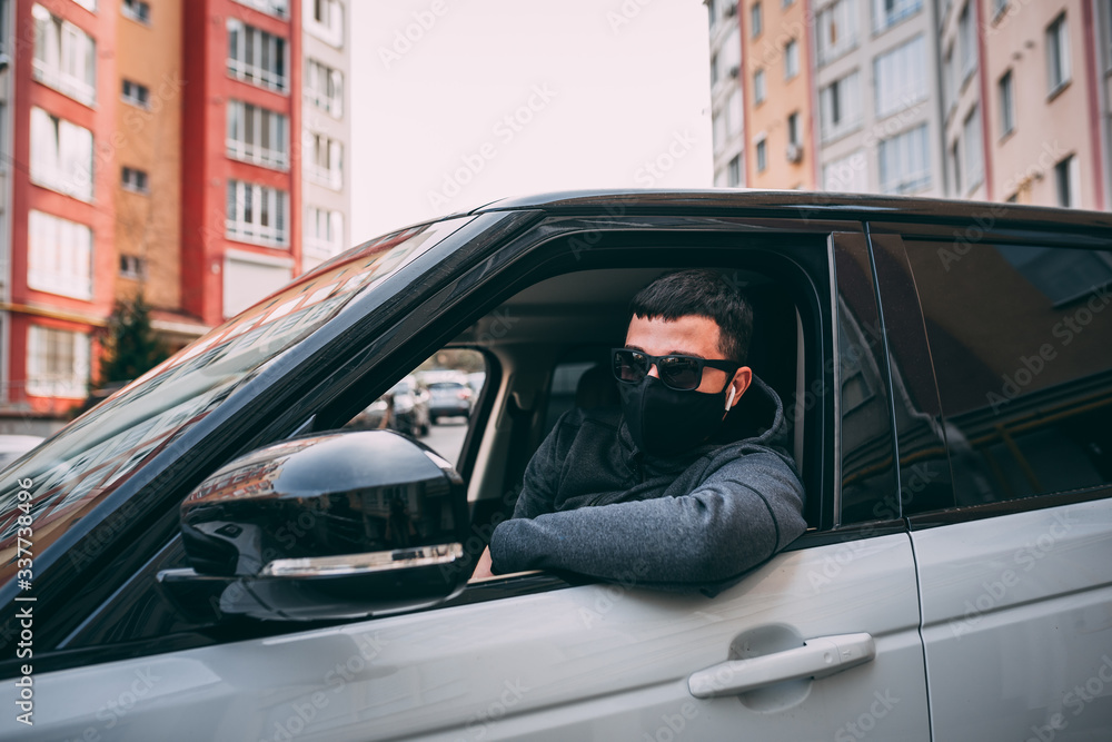 man in a black medical mask. handsome brunette driving a car, sitting at the wheel of an expensive car. thief, steal, coronavirus, disease, infection, quarantine, medical mask, covid-19