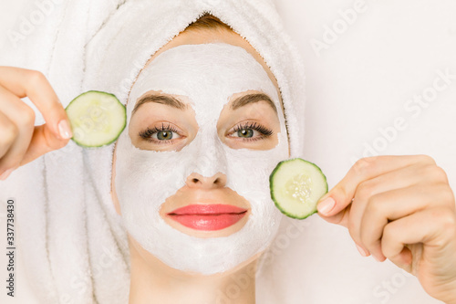 Home spa, skin care concept. Close up top portrait of pretty young woman with towel on her head, white clay mask on her face, holding pieces of fresh cucumber, lying on the isolated white background.