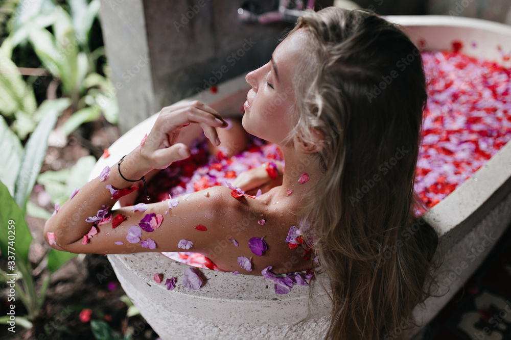 Obraz premium Long-haired tanned woman enjoying spa in weekend. Blonde female model lying in bath with flower petals and chilling with eyes closed.