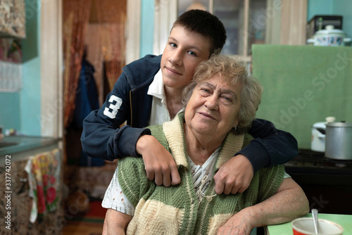 Teenager grandson hugs his grandmother in the kitchen at home. Poor, ordinary people.