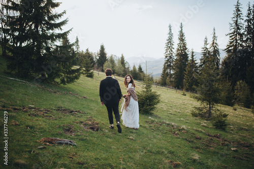 Fototapeta Naklejka Na Ścianę i Meble -  Wedding in the mountains. A young couple on a green slope among the pines, a girl in a white dress holds in her hands a bouquet of flowers and greenery with a ribbon