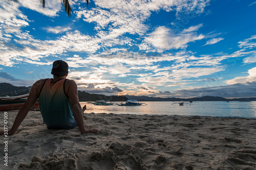 A young tourist man sitting on the sand of a beach in Port Burton while the sun sets in a cloudy day