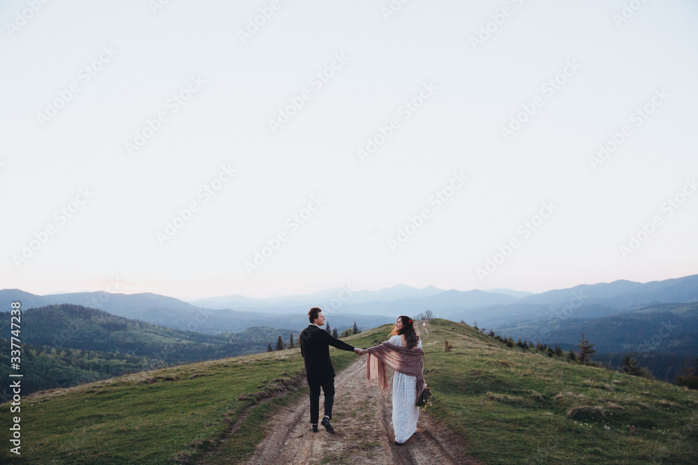 Wedding in the mountains. A guy and a girl in a white dress run along the road against the backdrop of mountains at sunset