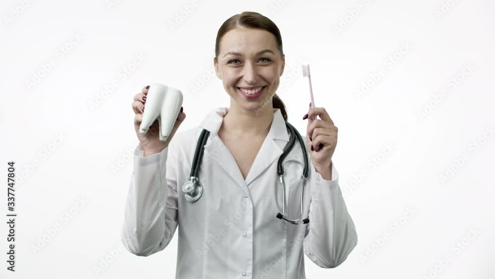 Young beautiful female dentist with perfect white smile in medical uniform with stethoscope holds healthy tooth layout and toothbrush. Oral care, dental health concept Young beautiful female dentist with perfect white smile in medical uniform with stethoscope holds healthy tooth layout and toothbrush. Oral care, dental health concept