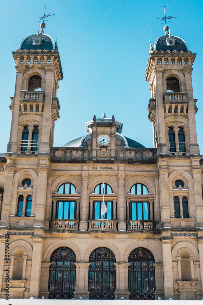 Town hall of the city of san sebastian in donostia, guipuzcoa, basque country, Spain, with two dark colored domes with pointed ends and a central clock. The sky is blue and cloudless. 