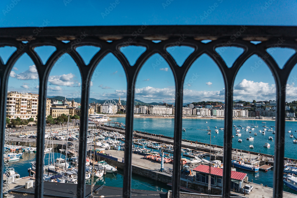 Historic center of the city of San Sebastián in Donostia, Guipúzcoa, Basque Country, Spain. La Concha beach seen from behind a black cast iron railing. The sky is blue and cloudy.