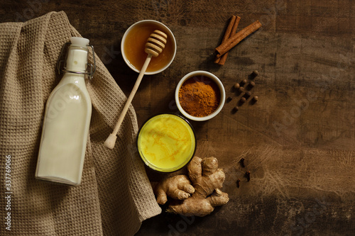 Turmeric Milk is poured into a glass. Next to it are the ingredients for making a drink - milk in a bottle on a napkin, spices and honey. Golden drink on dark moody background, selective focus. Top