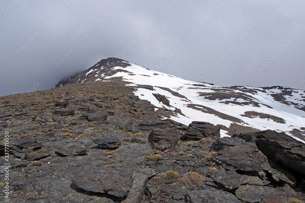 The mountain landscape with the dark rocks covered by white snow, the far peak, the cloudy sky.