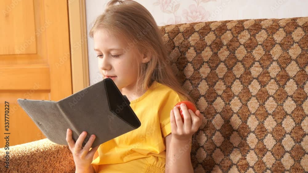 Distance learning. Home schooling. The girl is engaged on the tablet. Online training. The girl is sitting on the sofa, eating an Apple. Distance learning. Home schooling. The girl is engaged on the tablet. Online training. The girl is sitting on the sofa, eating an Apple.