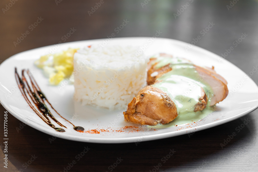 Chicken fillet with sauce is served with rice and vegetables on a white plate and a dark table. Close up. Chicken fillet with sauce is served with rice and vegetables on a white plate and a dark table. Close up.