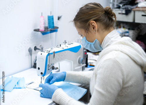 a girl in a protective mask sits at the workplace and sew sterile blue masks to protect against the virus. Sewing sterile masks