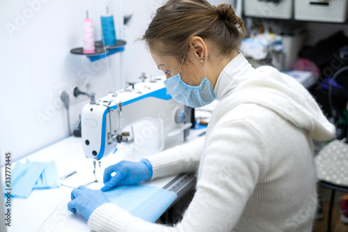 a girl in a protective mask sits at the workplace and sew sterile blue masks to protect against the virus. Sewing sterile masks