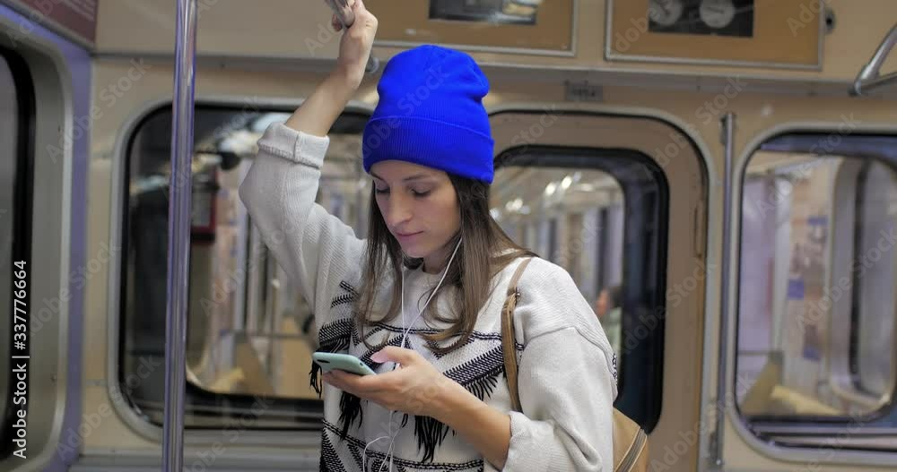Portrait of a cute girl in headphones and a blue hat on the subway, listening to music and browsing on a mobile phone in public transport. City lights background Portrait of a cute girl in headphones and a blue hat on the subway, listening to music and browsing on a mobile phone in public transport. City lights background