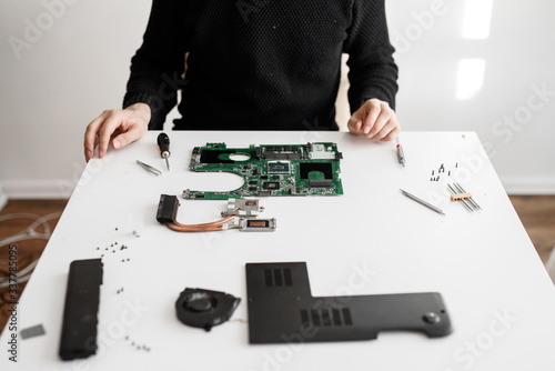 computer repairman sitting at a white table. on the table are different parts of a disassembled computer or laptop. Motherboard, cooling system, screws and screwdriver