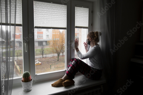 А young woman in a protective mask sits on the window sill. During the quarantine pandemic of the coronavirus covid-19. Russia.