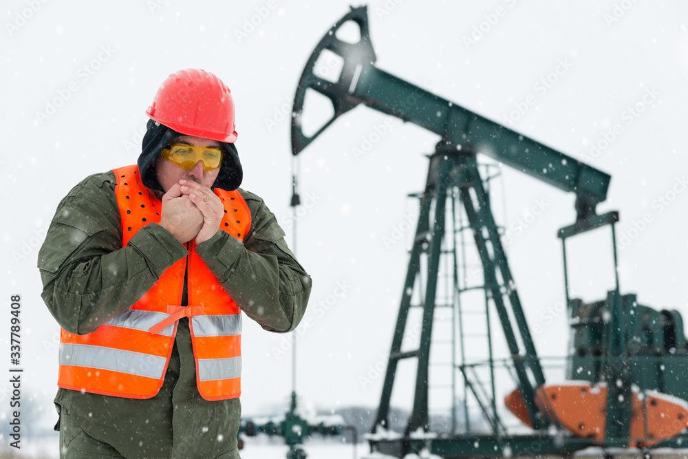 Oil Drill field , pump jack in the background .Oil and gas industry worker in safety clothing get it frozen in front , blowing hot air in his clasped hands