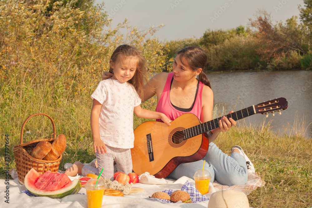 Mom and daughter play the guitar while sitting on the grass during a picnic in the park Mom and daughter play the guitar while sitting on the grass during a picnic in the park
