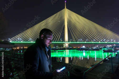 Young man looking at his phone in front of Ada bridge (most na Adi) in Belgrade, Serbia photo